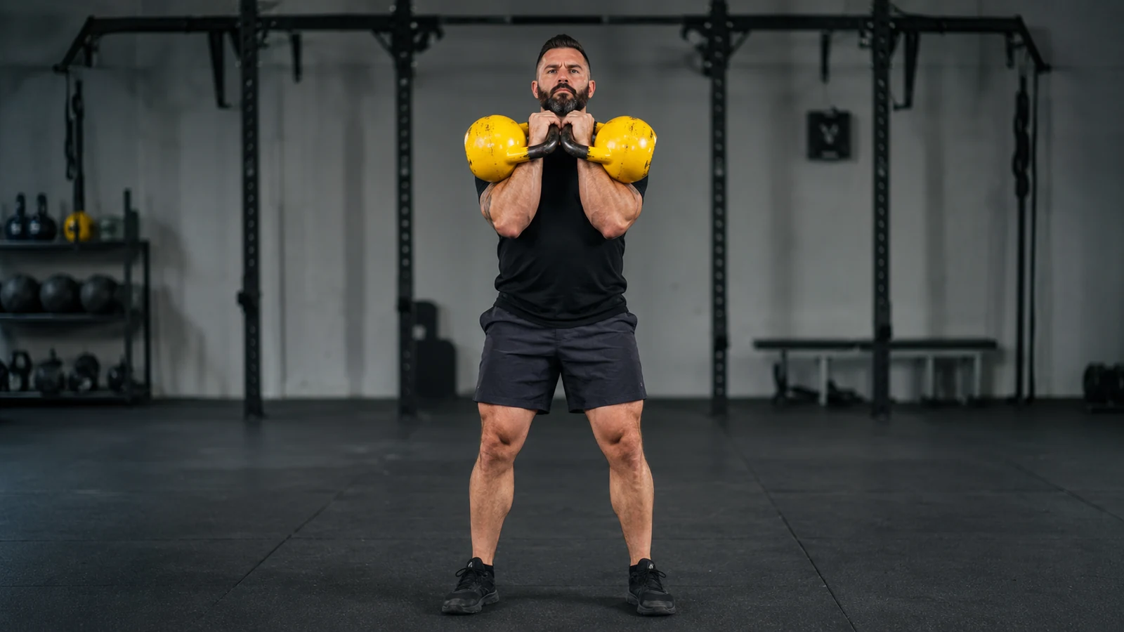 Kräftiger Athlet hält zwei gelbe Wettkampf-Kettlebells stabil in der Front-Rack-Position.